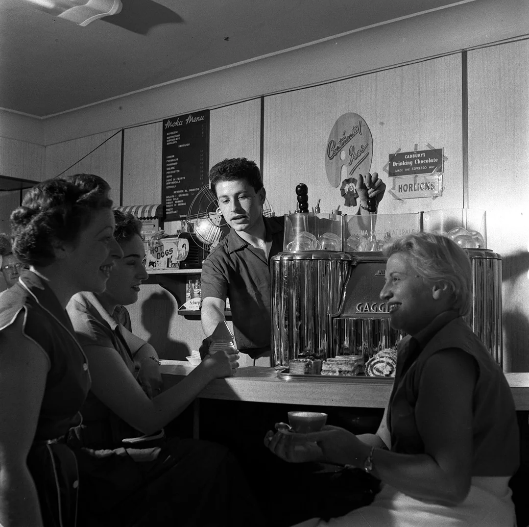A man serves three women at a vintage diner counter. The women are smiling and chatting, holding drinks. Menu boards and classic decor are visible in the background, creating a nostalgic, mid-20th-century atmosphere.