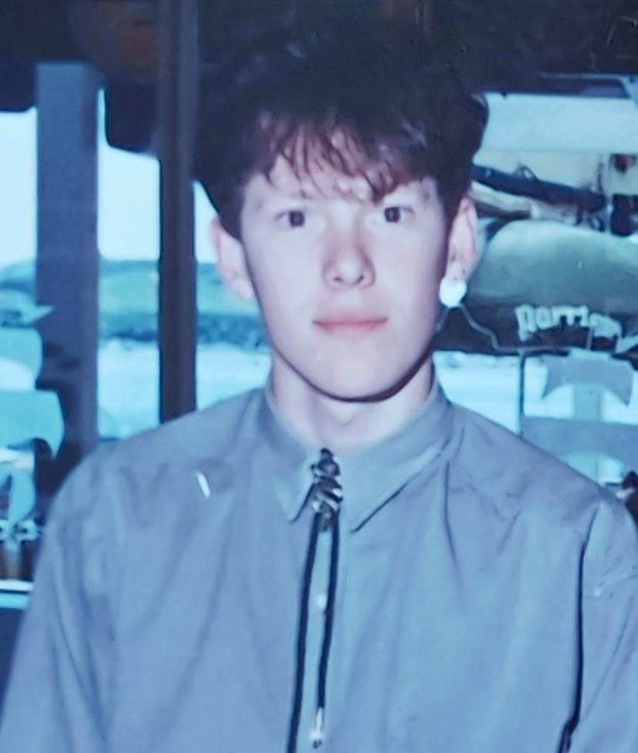 A young person with short brown hair wearing a gray shirt and bolo tie stands indoors. The background shows large windows with a view outside and some objects or signs.