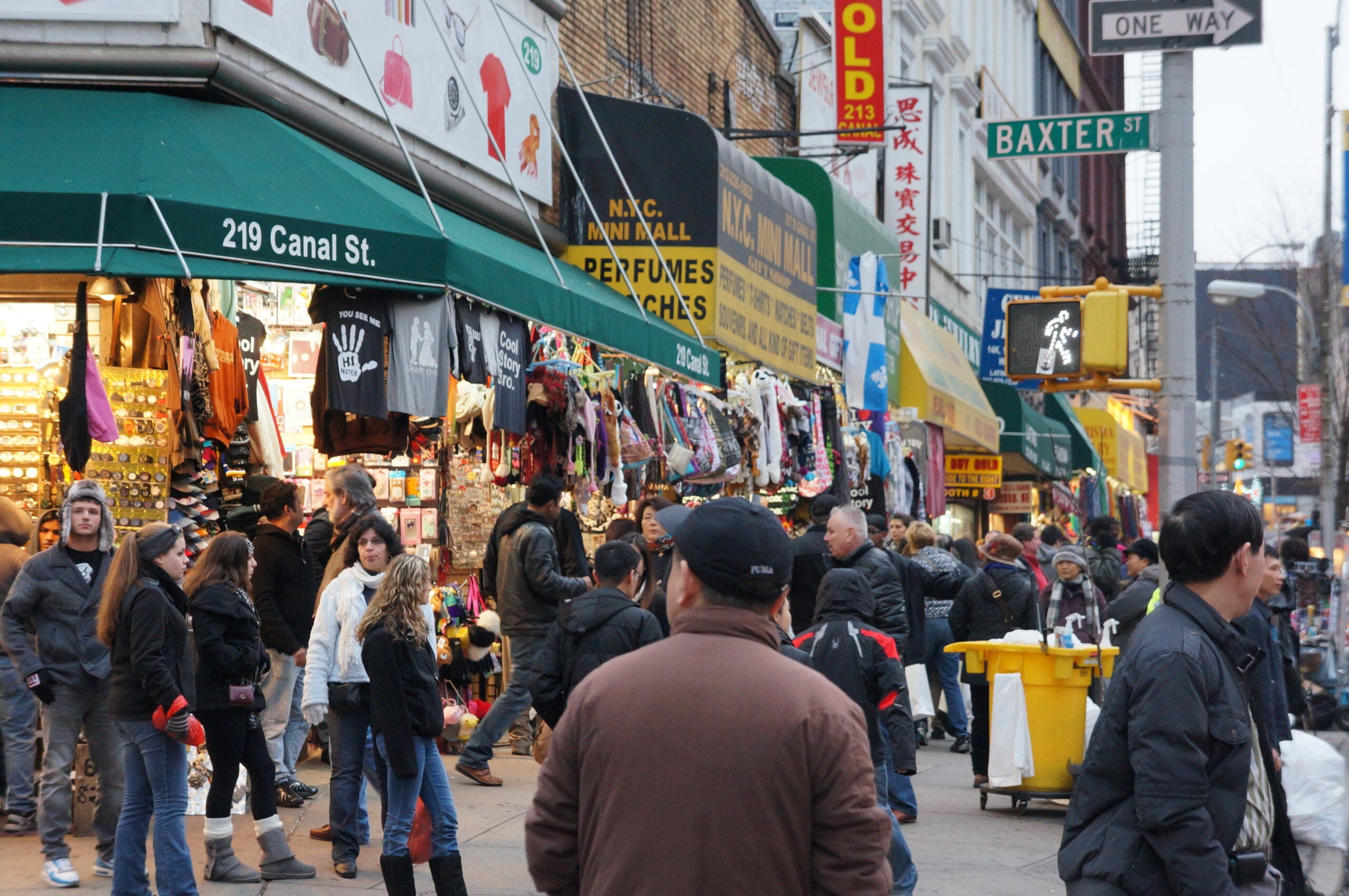 Busy street scene at Canal Street and Baxter Street in New York City, with people shopping, street vendors, colorful clothing displays, and storefronts under green awnings. It is daytime and pedestrians fill the sidewalk.