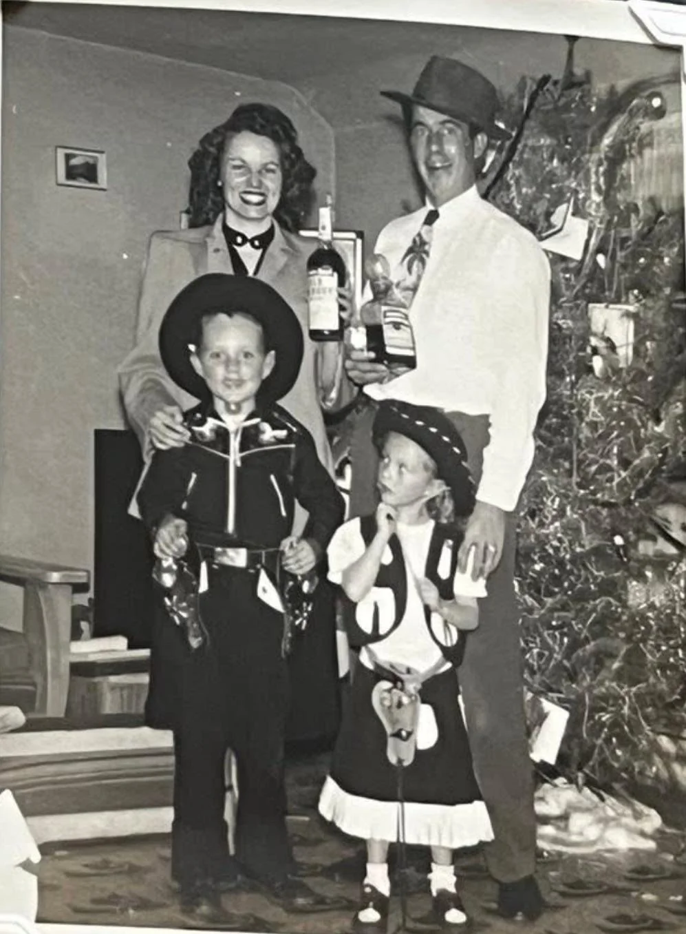 A black-and-white photo of a family of four posing indoors by a Christmas tree. The parents hold bottles and smile, while a boy and girl in cowboy costumes stand in front, the girl looking upward and touching her face.