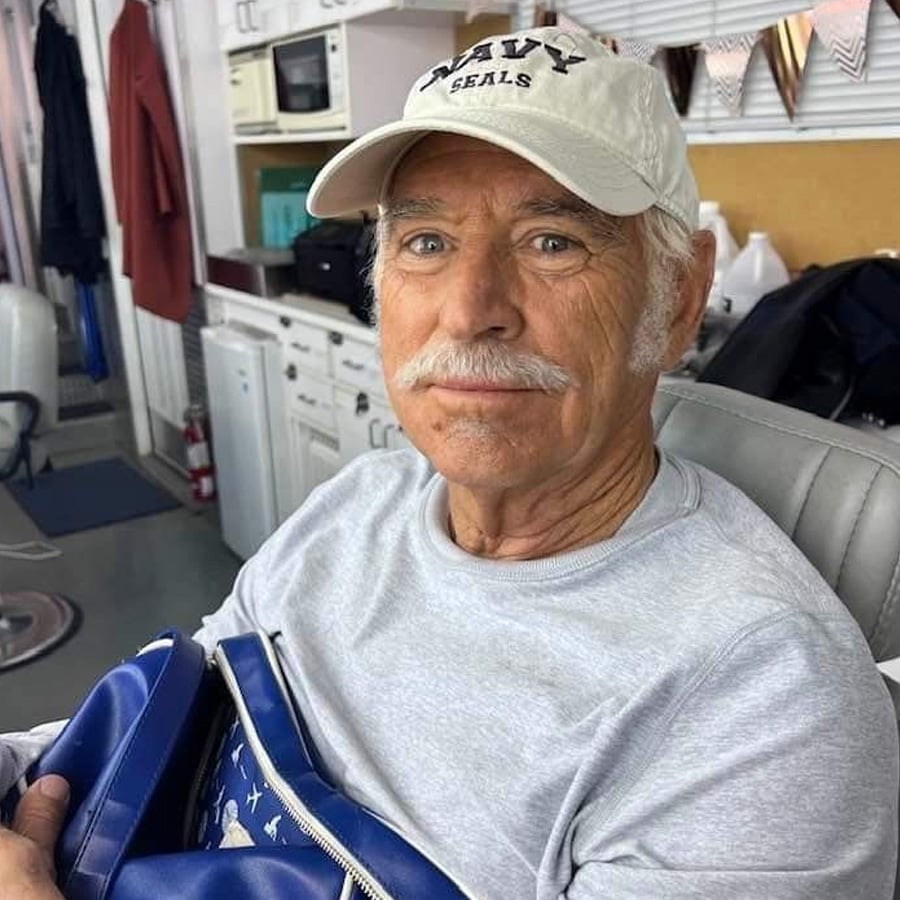 An older man with a white mustache and wearing a "NAVY SEALS" cap sits and smiles in a room with kitchen cabinets and decorations.
