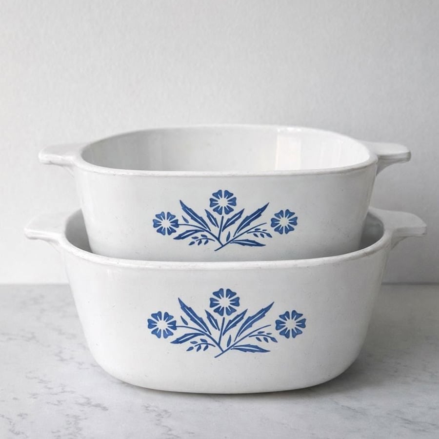 Two white vintage casserole dishes with blue floral patterns are stacked on a white surface, against a plain light background.