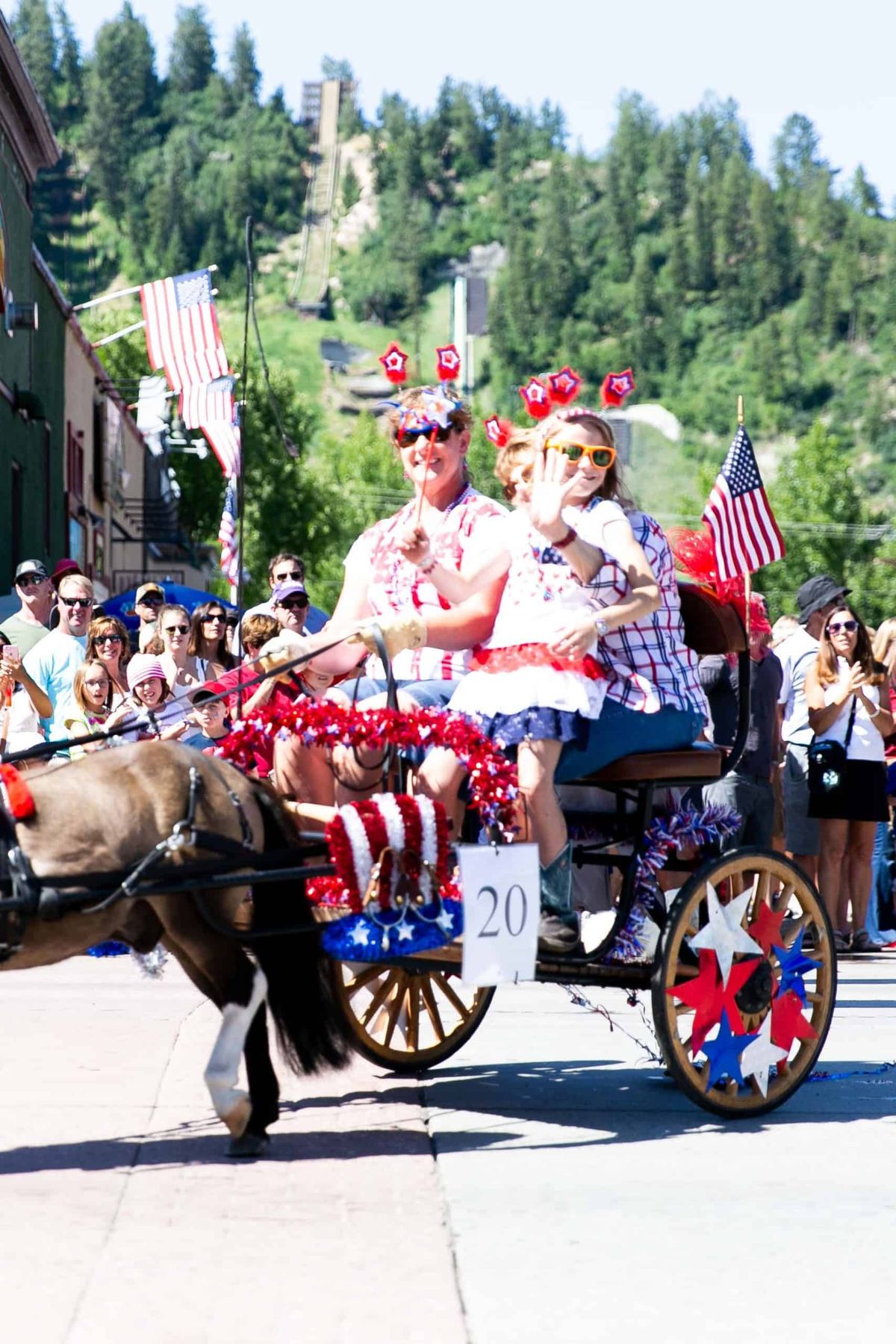 Two people dressed in patriotic attire ride a decorated cart pulled by a miniature horse in a parade. The cart has American flags and festive decor. Spectators line the street, and green hills are visible in the background.