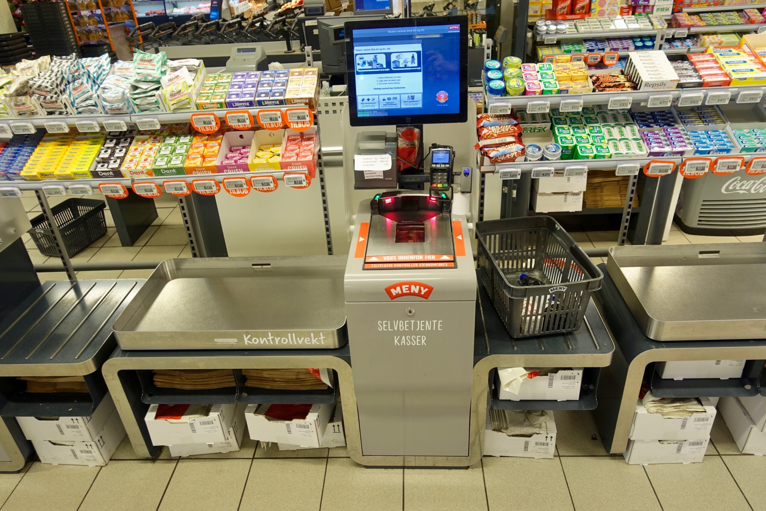 A self-checkout station at a grocery store, with a basket on the counter, scales, a barcode scanner, shelves of products in the background, and a screen displaying instructions above the scanner.