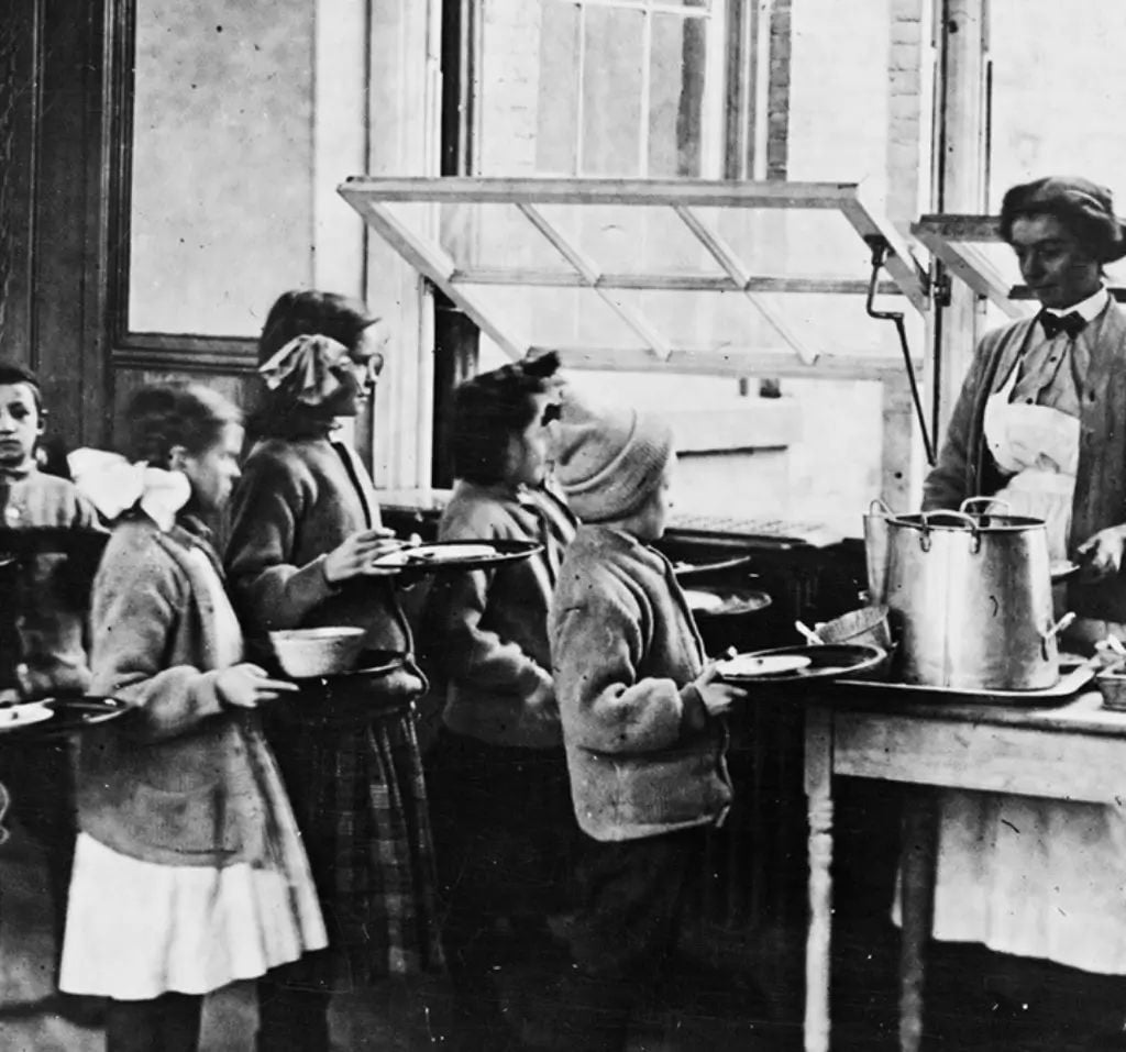 Children in line at a cafeteria, holding trays, while a woman in an apron serves food from a large pot by a window; black and white historical photo.