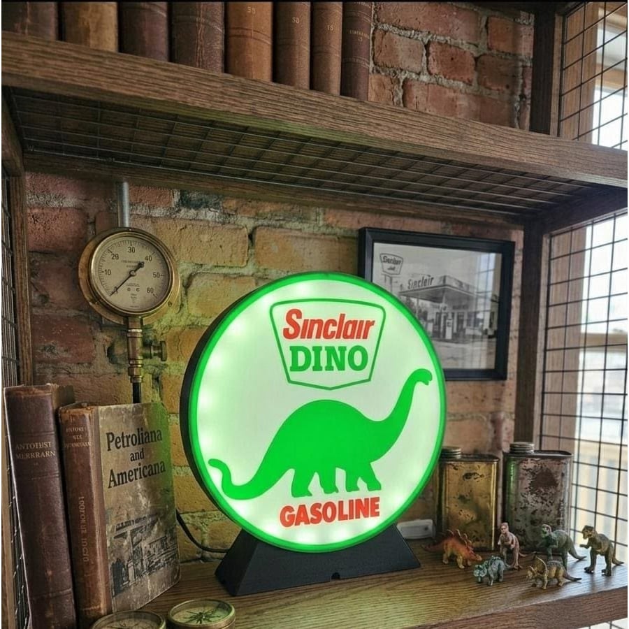 A lit Sinclair Dino Gasoline sign with a green dinosaur sits on a shelf next to vintage books, a pressure gauge, toy dinosaurs, and a framed black-and-white photo against a brick wall.