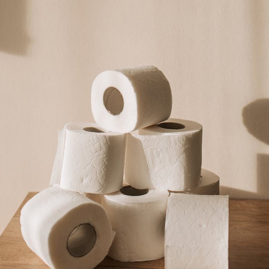 Seven rolls of white toilet paper, some stacked and some lying on their sides, are arranged on a wooden surface against a beige background in soft, natural light.