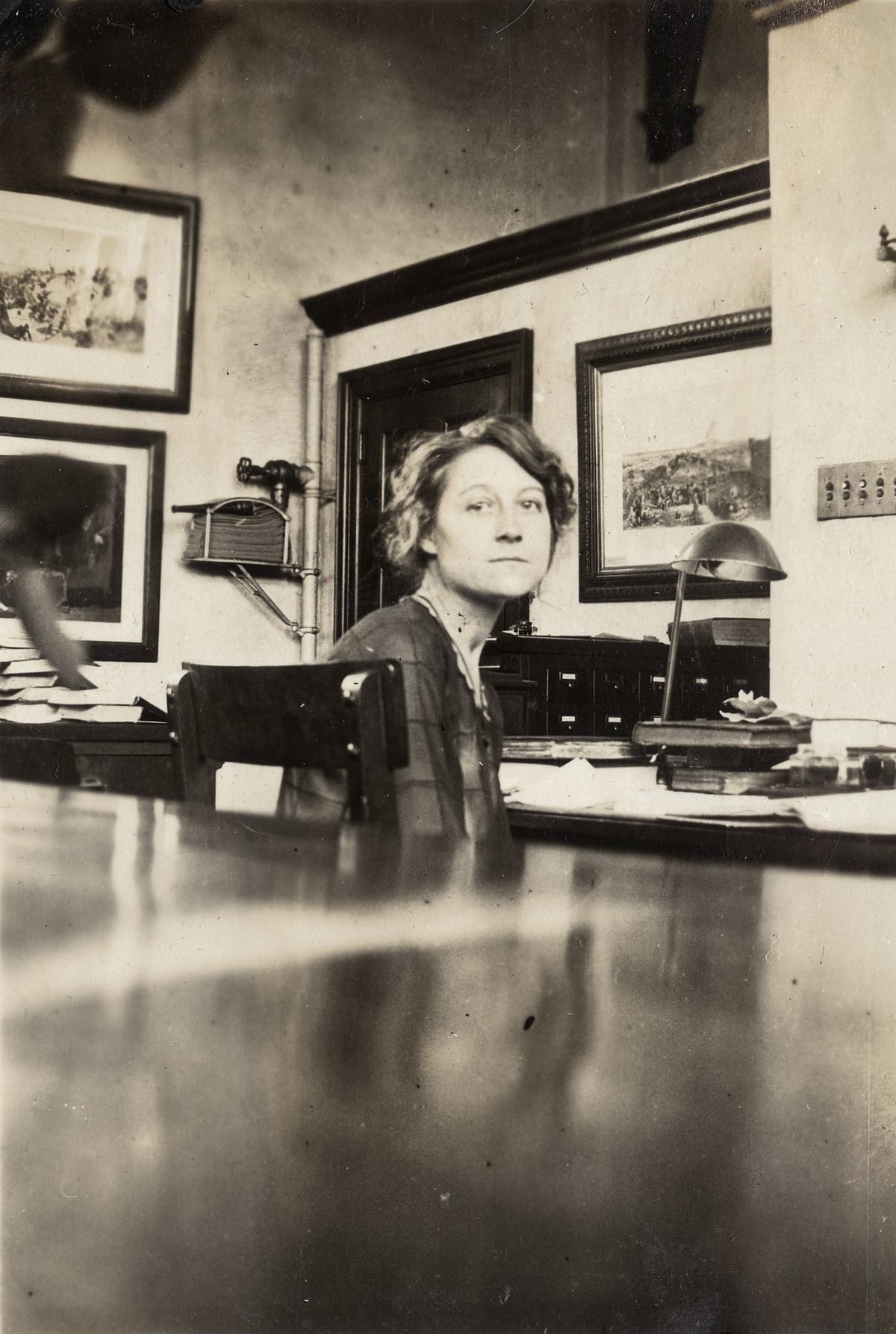 Black-and-white photo of a woman sitting at a desk in an office, looking at the camera. There are framed pictures on the wall, a lamp, and paperwork on her desk.