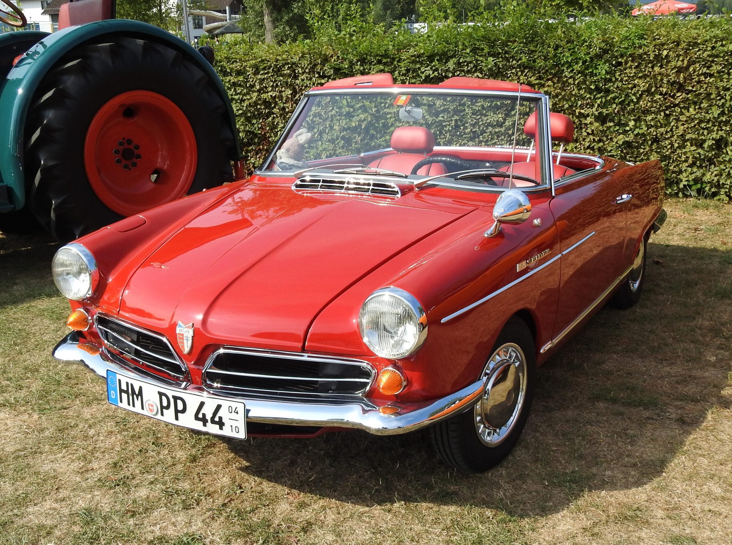 A shiny red vintage convertible car with chrome details is parked on grass near a hedge, with part of a large green vehicle visible in the background.