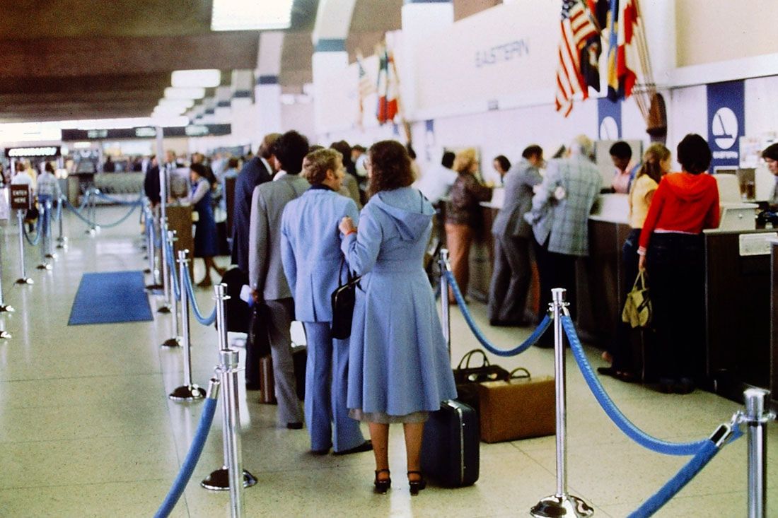 People wait in line at an airport check-in counter. Most are dressed in 1970s-style clothing. Blue rope barriers guide the line, and several suitcases are visible. Flags hang above the counters in the background.