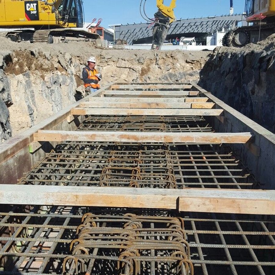 A construction worker in safety gear stands in a trench with steel reinforcement bars and wooden supports. Excavators and construction equipment are visible in the background.