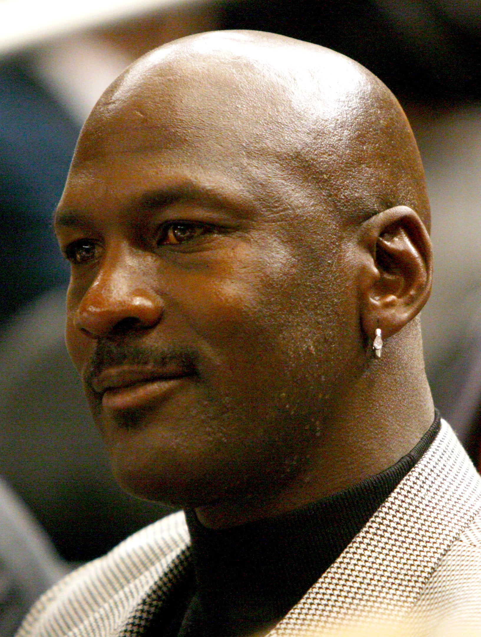 A man with a shaved head and mustache wears a diamond earring and a checked blazer over a black shirt, looking slightly to the side with a calm expression.