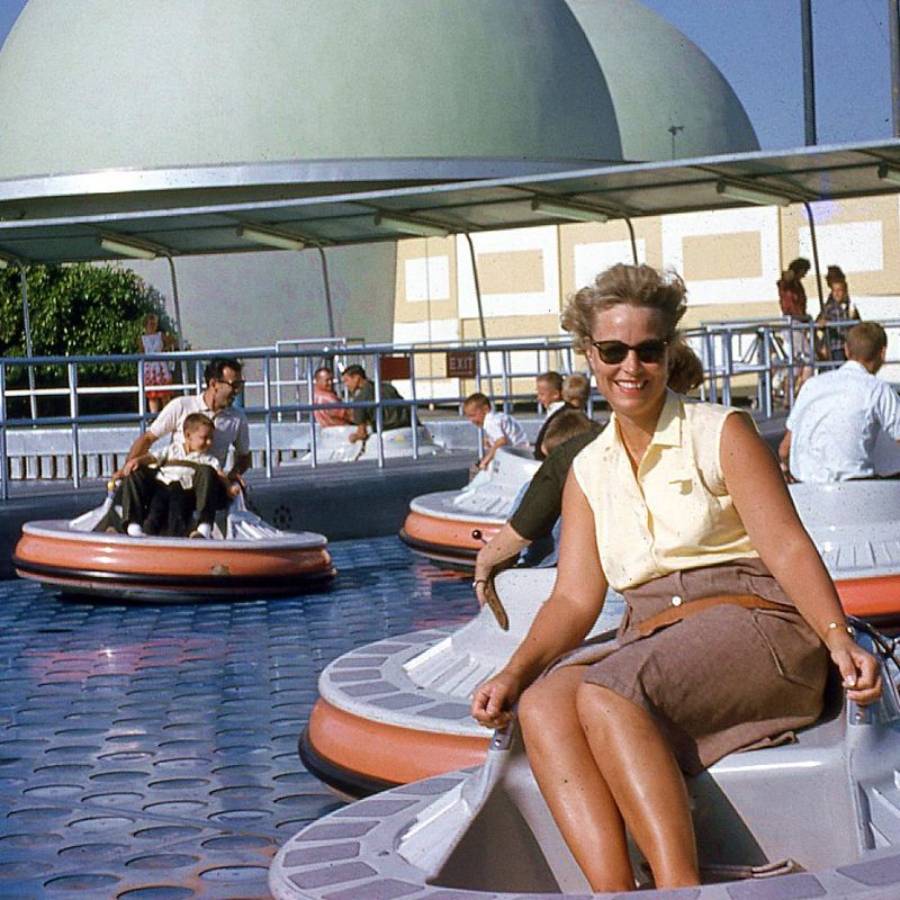 A smiling woman in sunglasses sits on a round bumper car ride outdoors, with other people enjoying the ride in the background. The scene is sunny, and a large dome structure is visible behind the ride.