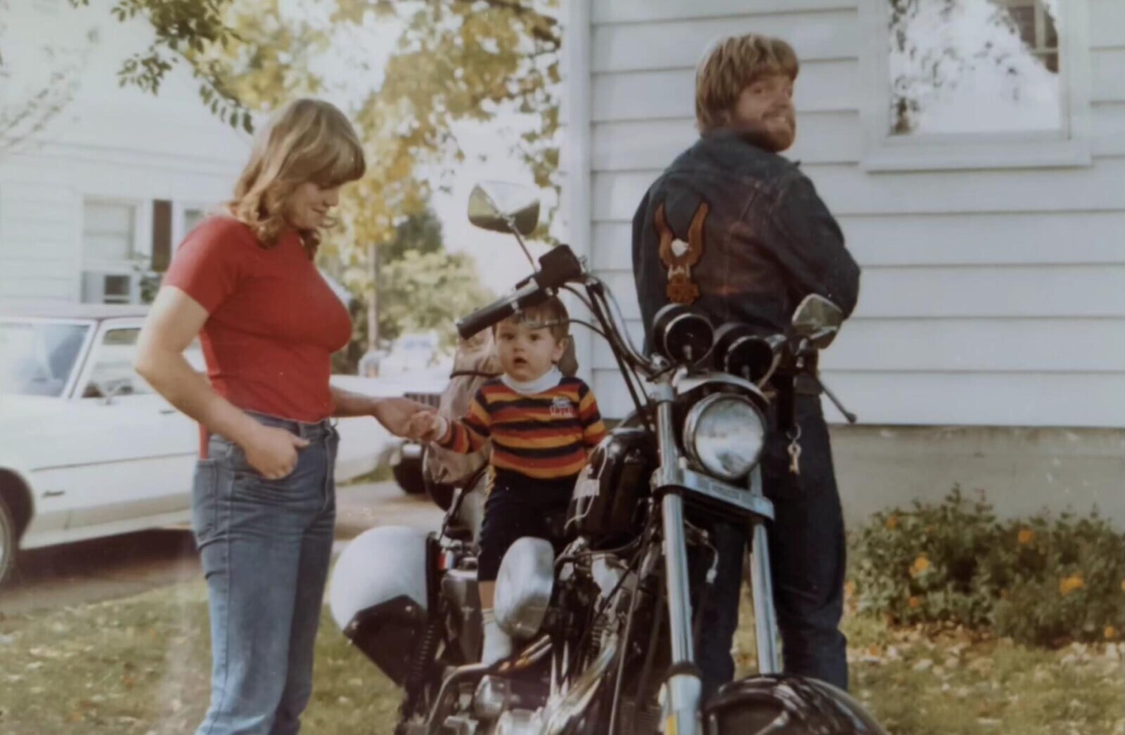 A woman stands beside a man and child sitting on a motorcycle. The man wears a denim jacket with an eagle patch and looks back, while the child, in a striped sweater, sits on the motorcycle. A white house is in the background.