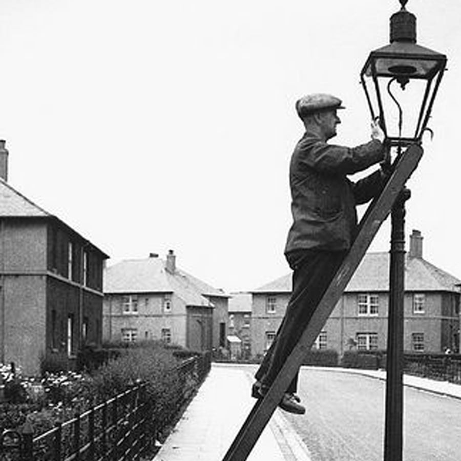A man in a flat cap stands on a ladder, lighting or repairing a streetlamp in a residential neighborhood lined with houses.