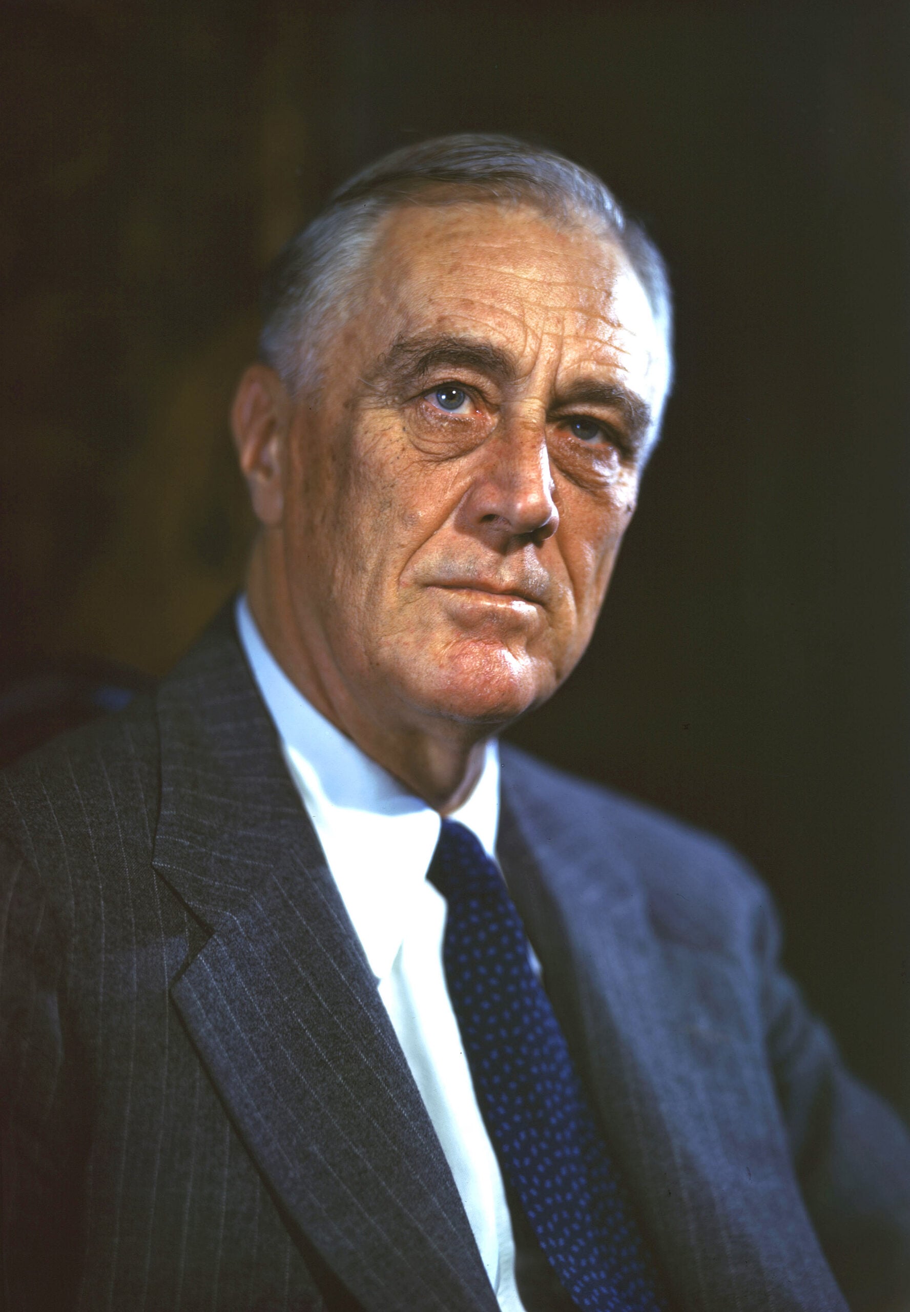 A mature man in a suit and blue tie looks slightly past the camera with a serious expression, set against a dark background.