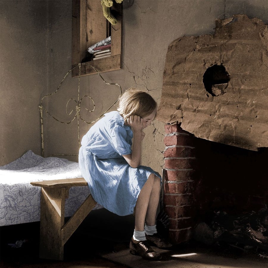 A young girl in a blue dress sits on a bench by a bed, leaning her head on her hand, next to a damaged brick fireplace in a dimly lit, sparsely furnished room.