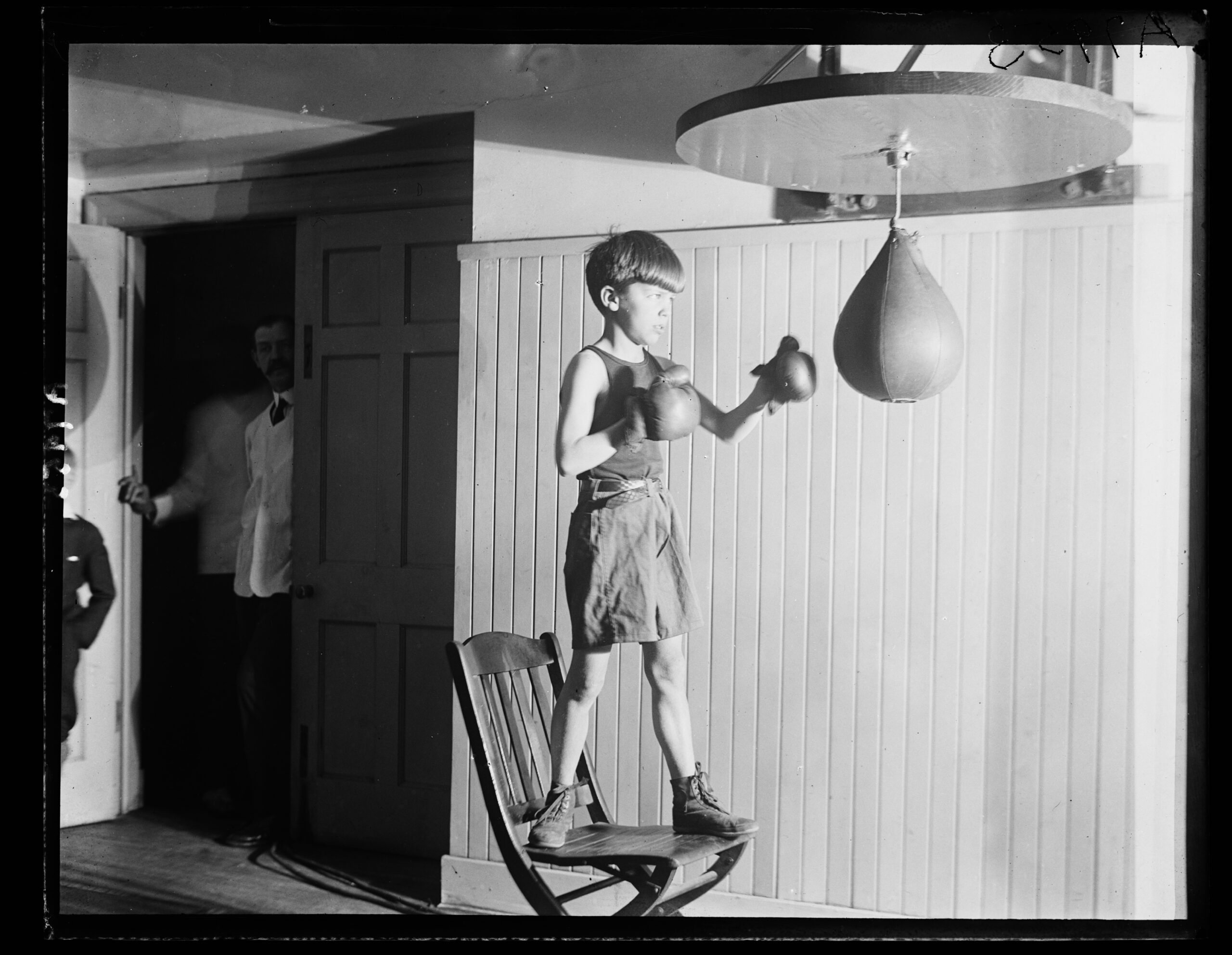 A young boy stands on a chair wearing boxing gloves and shorts, punching a speed bag in a room with paneled walls. A man watches from the doorway in the background.