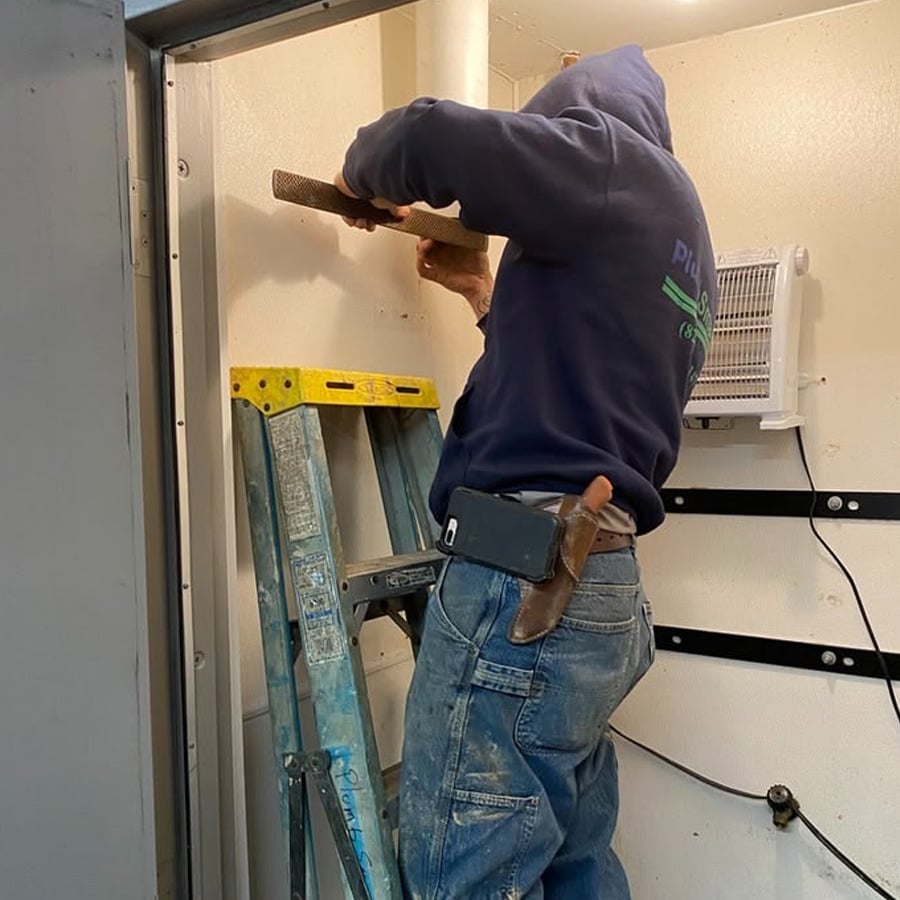A person in a blue hoodie stands on a step ladder, using a tool on a white wall. They have a phone and a holstered tool on their belt.