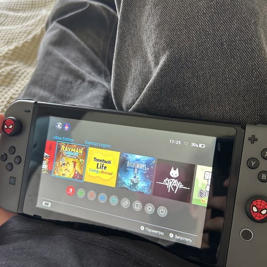 A person holding a Nintendo Switch with Spider-Man joystick covers, displaying the home screen with games like Rayman Legends, Stray, and The Nightmares.