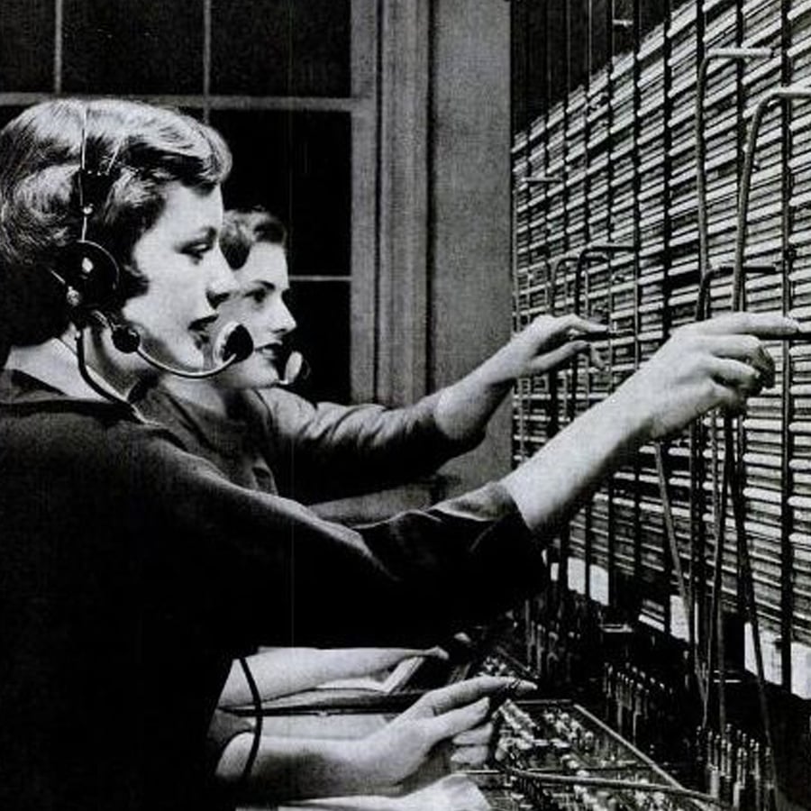 Two women wearing headsets operate a vintage telephone switchboard, connecting calls by plugging cables into a large panel filled with sockets and wires.