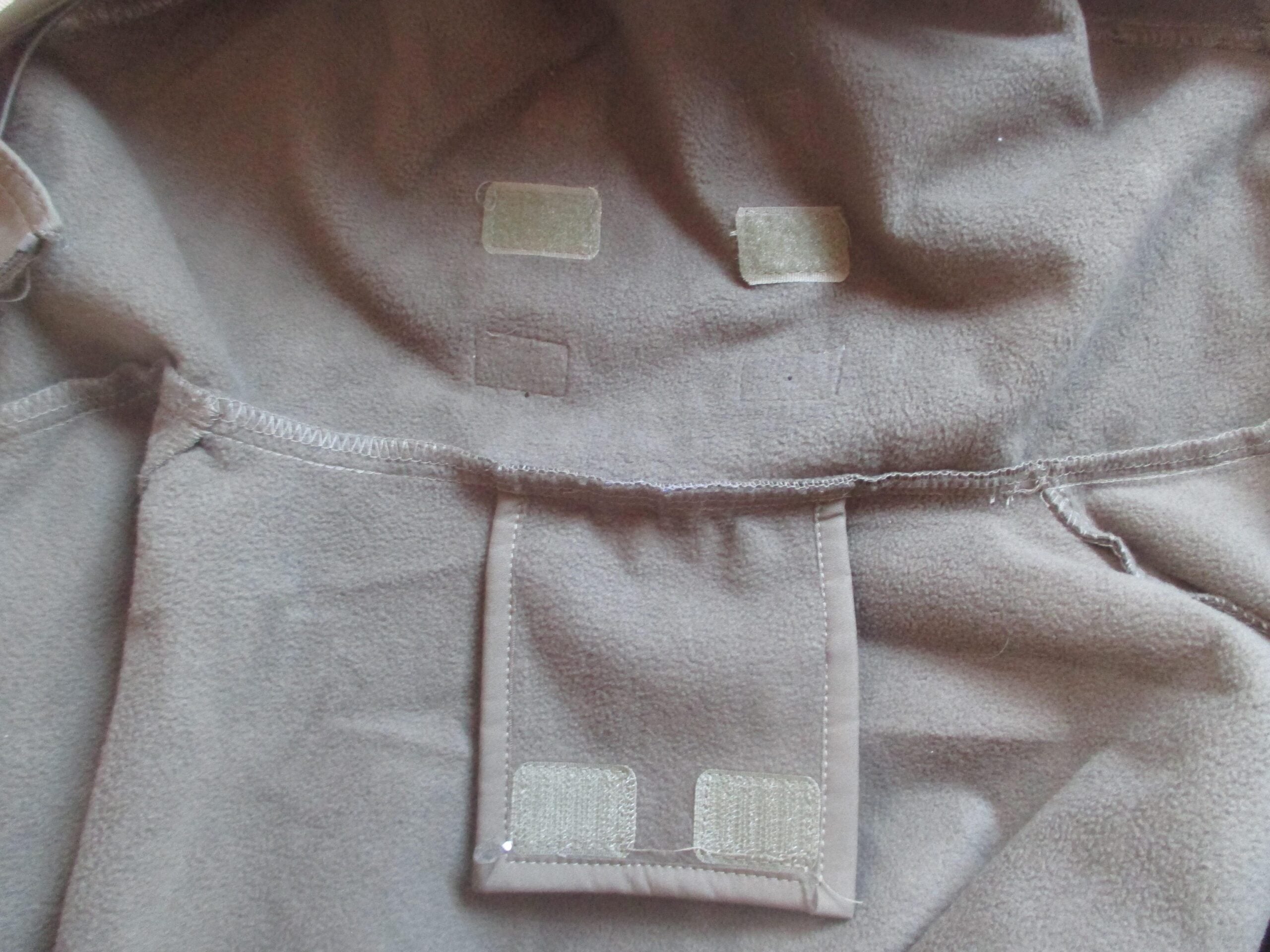Inside view of a brown fleece jacket showing three rectangular Velcro patches sewn on the upper back and one large rectangular flap with two Velcro patches at the bottom.