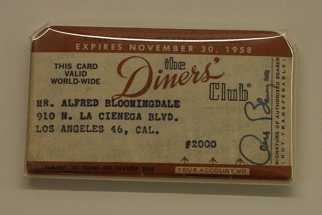A vintage Diners Club card with the name Alfred Bloomingdale, an expiration date of November 30, 1958, and an address in Los Angeles, CA. The card is enclosed in plastic.