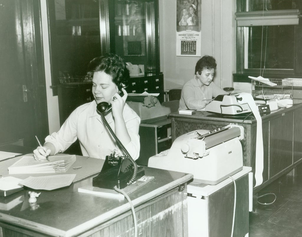 Two women work in an office; one speaks on a rotary phone and writes notes, while the other types on a typewriter at a desk. Office furniture, papers, and shelves are visible in the background.