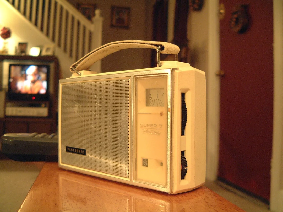 A vintage white Panasonic Super 7 radio sits on a table in a warmly lit living room, with a TV and staircase visible in the background.
