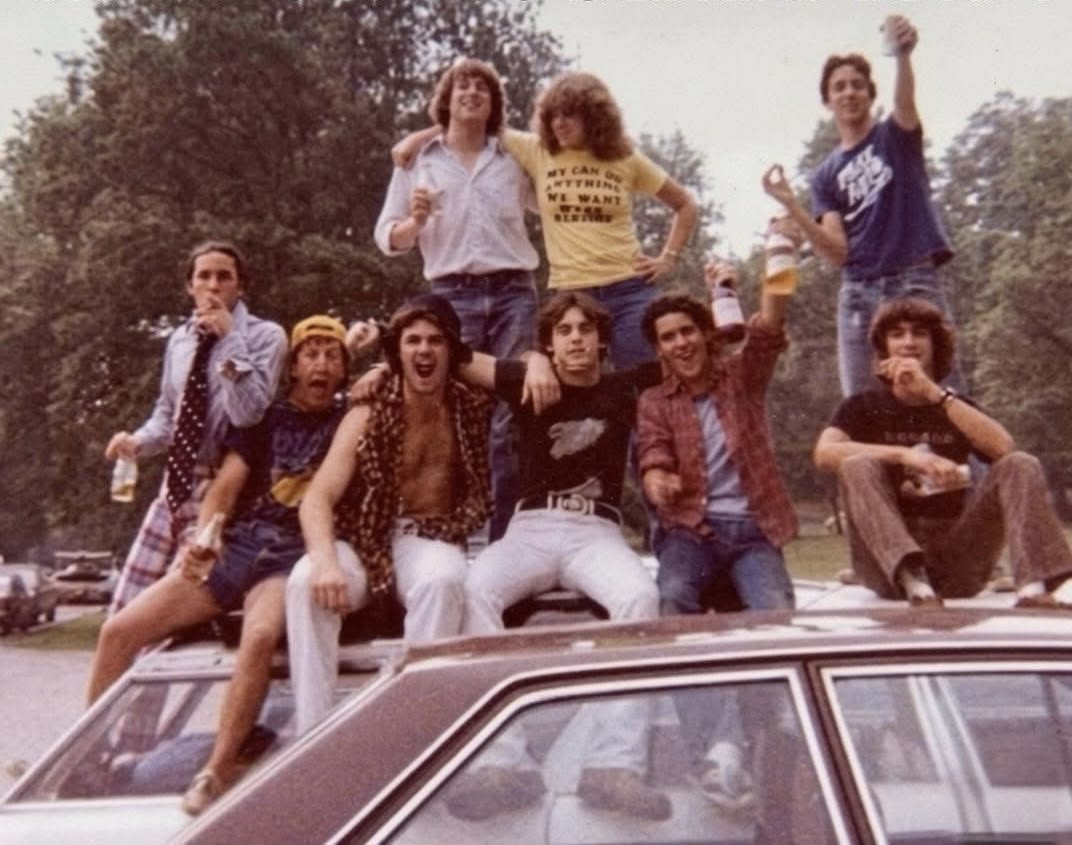 A group of young people in casual 1970s clothes pose and smile on top of a car in a park, some holding drinks. Trees and grass are visible in the background.
