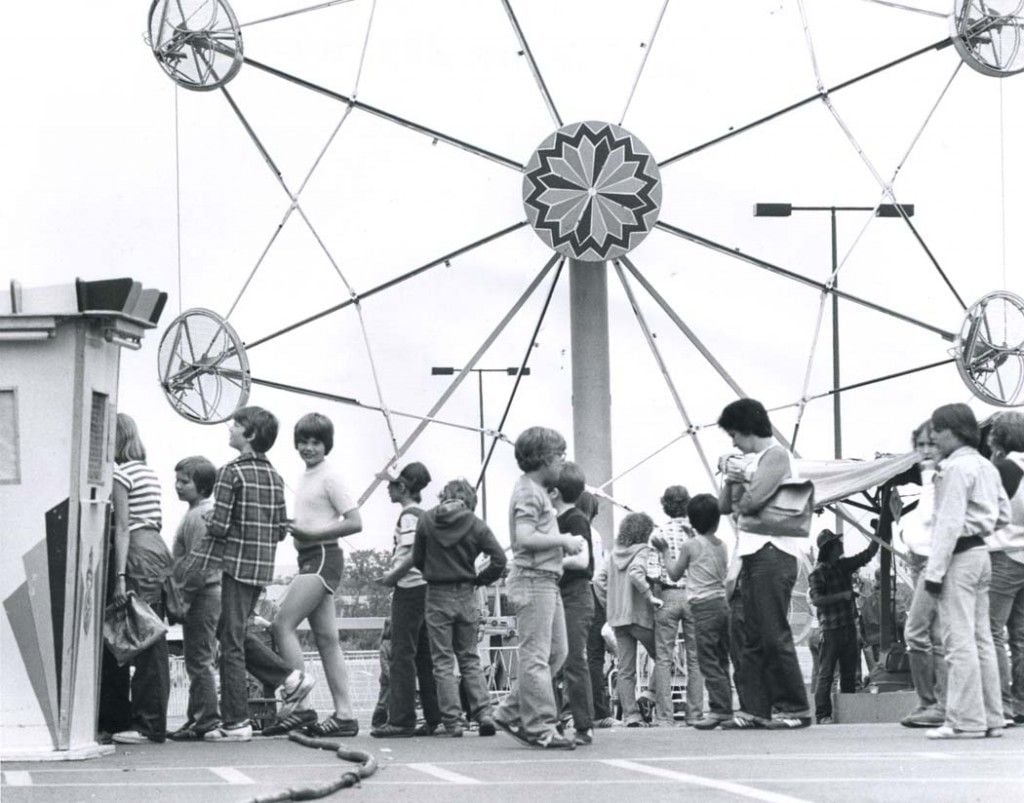 A group of children and teenagers wait in line at a carnival in front of a Ferris wheel. Some look at the camera, while others face forward or talk. The scene is outdoors, and the image is in black and white.