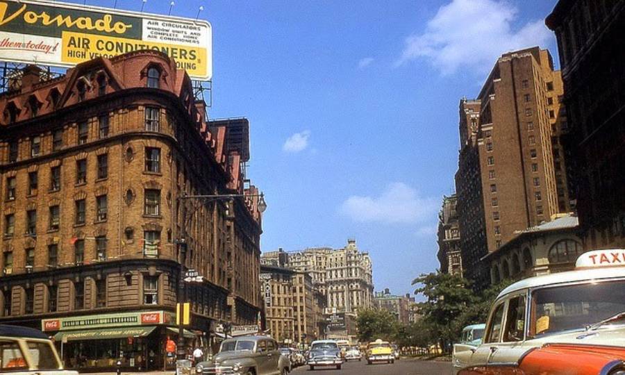 A busy city street scene from the mid-20th century with vintage cars, tall brownstone buildings, a taxi in the foreground, and a large air conditioner billboard against a blue sky.