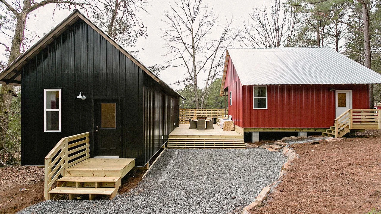 Two small houses, one black and one red, with wood porches and steps, sit among trees. A gravel path leads to the houses, and outdoor seating is arranged on the connecting deck.