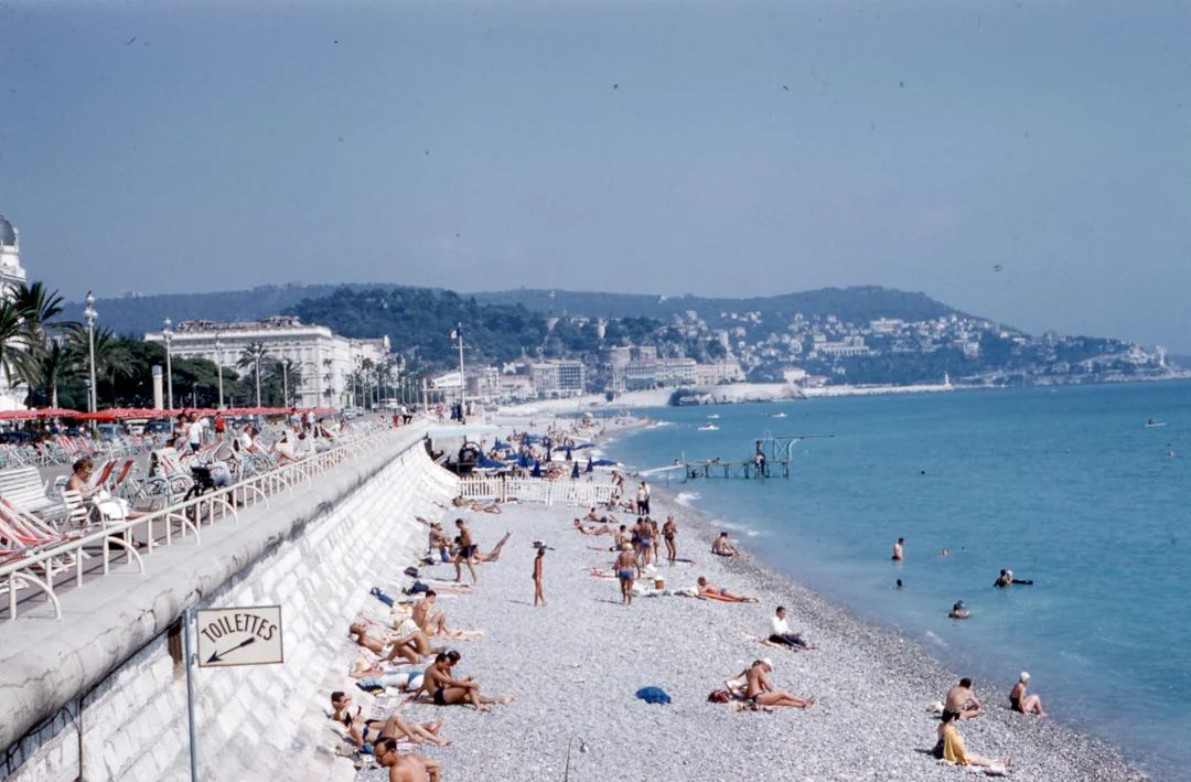 People relax on a pebbly beach by a blue sea, with a promenade, palm trees, and distant hills in the background. A "Toilettes" sign is visible along the walkway. The sky is clear and sunny.