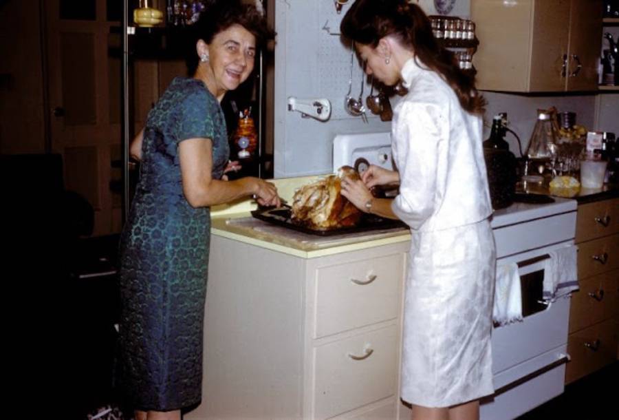 Two women carve a roasted turkey in a vintage kitchen. One woman smiles at the camera while the other focuses on cutting the meat. Shelves and a stove are visible in the background.