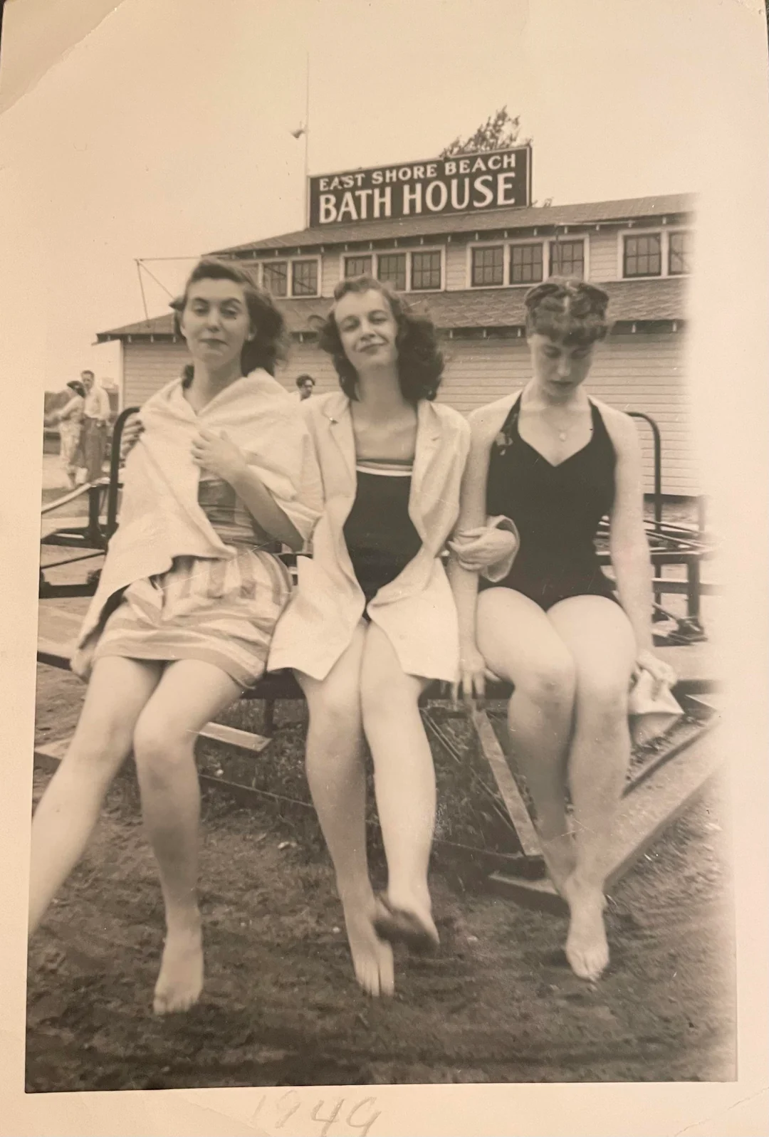 Three women in swimsuits sit on a bench, smiling, in front of a building with a sign reading "East Shore Beach Bath House." The photo is black and white and appears vintage.
