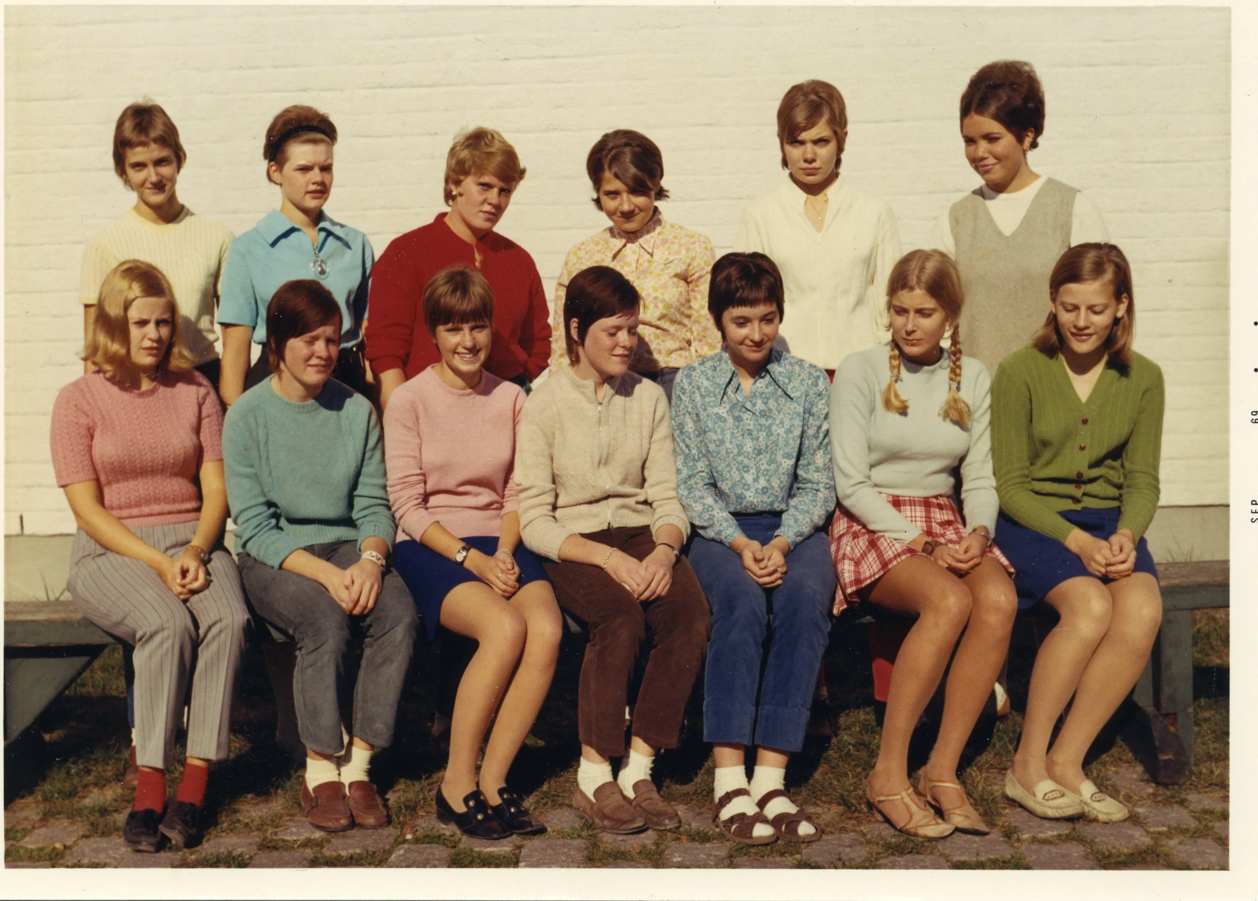 A vintage photo of thirteen young women posing in two rows outside against a white wall. The front row sits on a bench, and the back row stands. They wear colorful sweaters and skirts or pants, with a mix of short and long hairstyles.