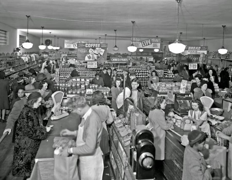 Black-and-white photo of a busy grocery store in the mid-1900s, with shoppers filling carts, cashiers at registers, and shelves stocked with canned goods, coffee, bread, and other groceries under hanging light fixtures.