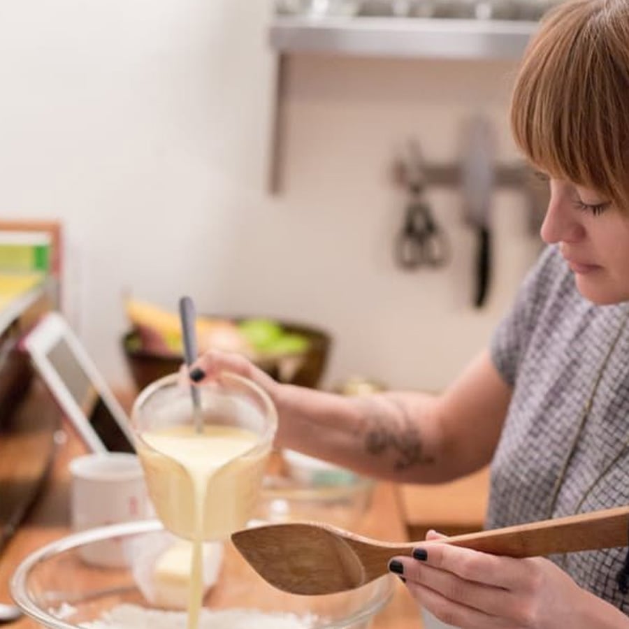 A person pours liquid batter from a measuring cup into a mixing bowl while holding a wooden spoon, preparing ingredients in a kitchen.