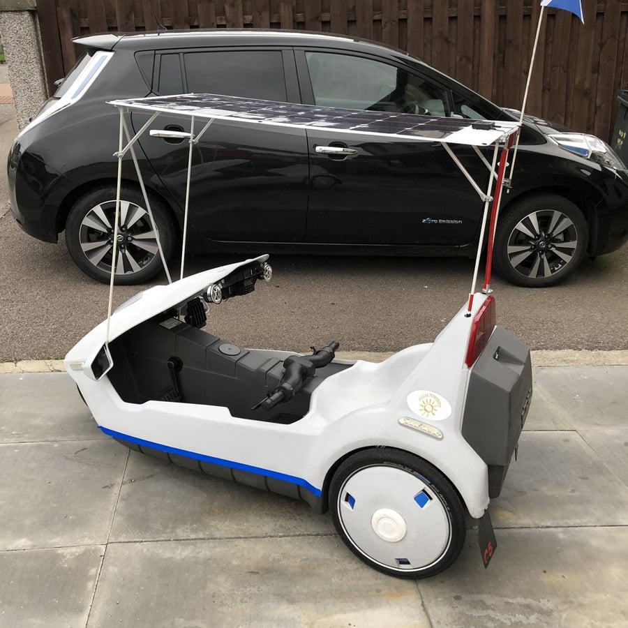 A small, white, single-seat electric vehicle with a solar panel roof is parked on a driveway. A black car is parked on the street behind it, next to a wooden fence.