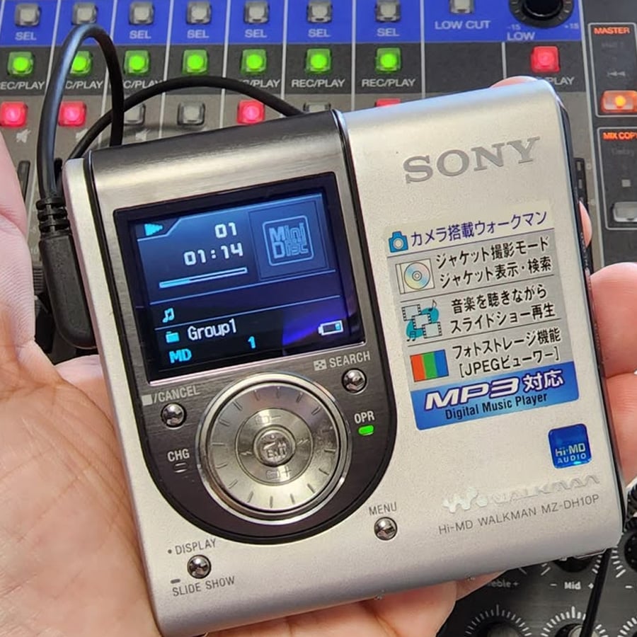 A hand holds a silver Sony Hi-MD Walkman MP3 player, displaying track info on a small screen. Colorful mixer controls are blurred in the background.