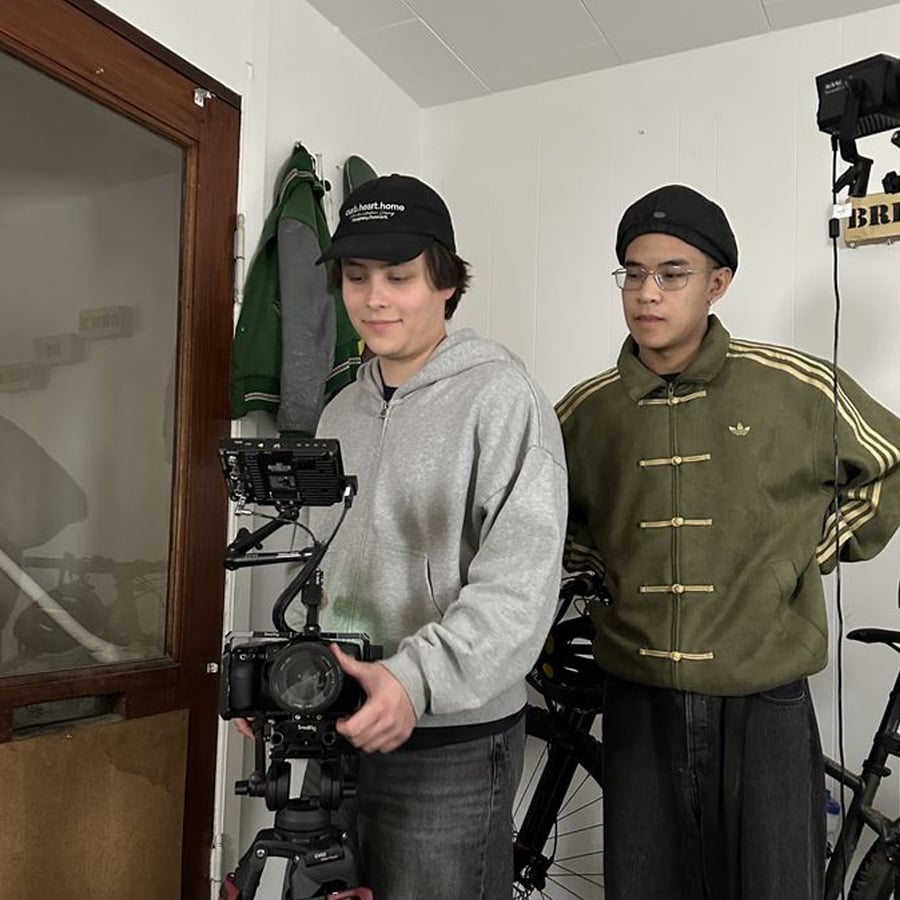 Two people stand indoors near bicycles. One, wearing a gray hoodie and cap, operates a camera on a tripod, while the other, in an olive jacket and glasses, observes. They appear focused and are in a small, cozy room.