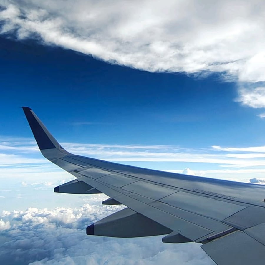 View from an airplane window showing a wing and winglet with blue sky and clouds above and below.