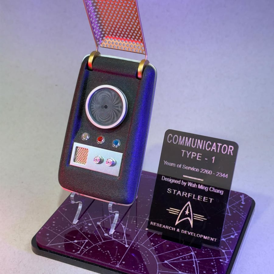 A Star Trek communicator prop on a display stand, next to a plaque with details: "Communicator Type-1," years of service, designer name, and Starfleet Research & Development logo.
