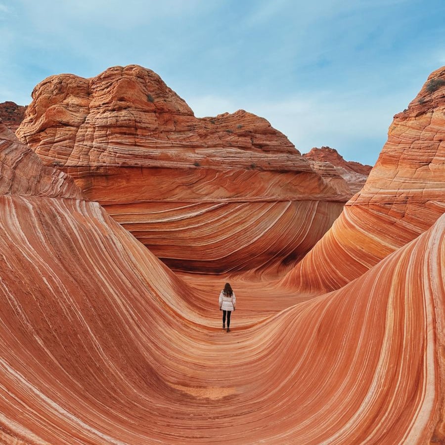 A person stands in the middle of a sandstone formation with dramatic orange and white swirling stripes, under a blue sky.