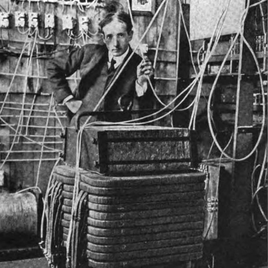A person in a suit stands with one hand on their hip, holding an object, surrounded by wires and electrical equipment in a workshop or laboratory setting.