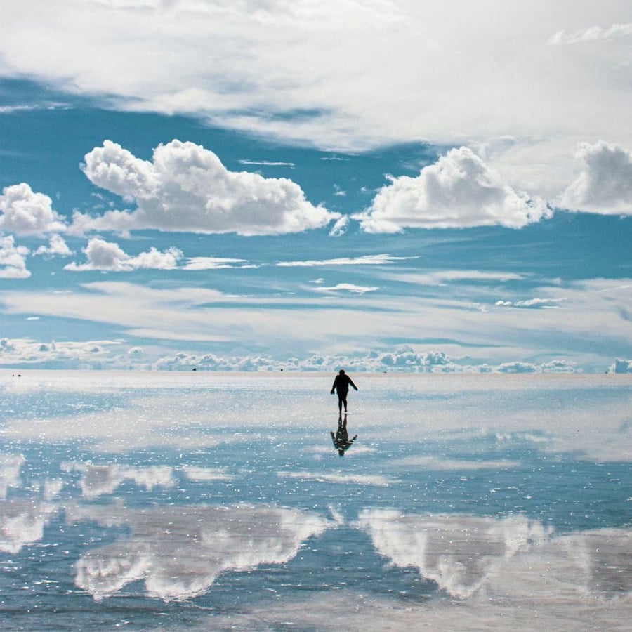 A person walks alone on a vast, reflective surface under a blue sky with scattered clouds, creating a mirror-like illusion with the clouds reflected below.