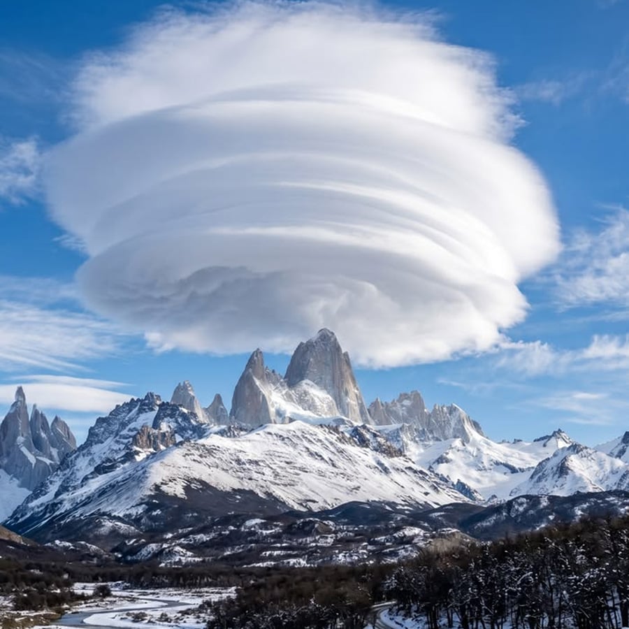 A large, layered lenticular cloud hovers above a snow-covered mountain range under a blue sky, creating a dramatic and surreal scene.