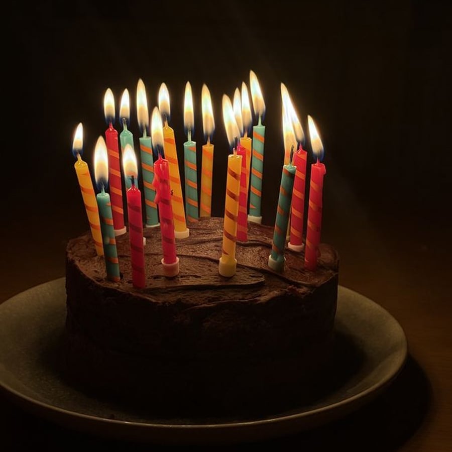 A chocolate birthday cake on a plate with 20 colorful lit candles, glowing brightly in a dark room.