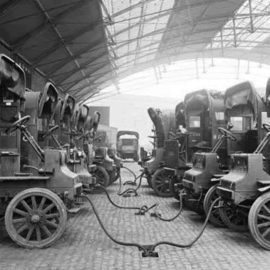 Several early 20th-century electric vehicles are charging under a large arched roof, connected by thick cables on a cobblestone surface. The vehicles are lined up in two parallel rows.